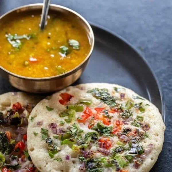 Two Sri Balaji uttapam with vegetables and a bowl of sambar on a dark plate.