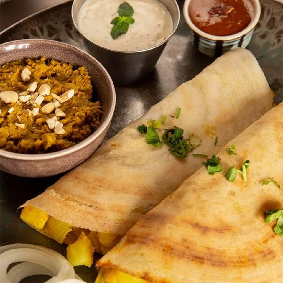 Two golden-brown Sri Balaji dosas filled with potatoes, served with chutney, sambar, and onions.